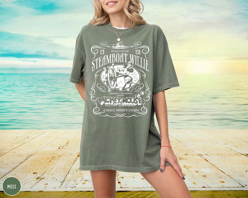May include: An olive green t-shirt featuring a vintage design with the text "Steamboat Willie" and "Mickey Mouse". The graphic includes an illustration of Mickey Mouse and a steamboat. The shirt is a relaxed fit.