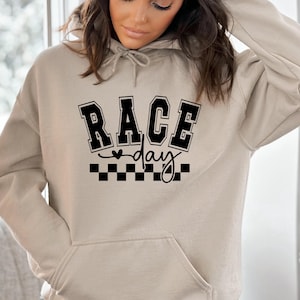 May include: A beige hoodie with a front pocket and drawstring hood. The hoodie features the text "RACE day" in a black, collegiate-style font above a black and white checkered flag design.