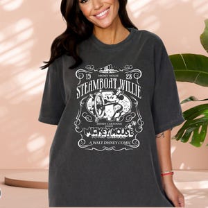 May include: Dark gray t-shirt featuring a white graphic of Mickey Mouse from the 1928 cartoon "Steamboat Willie." The design includes the text "Disney Cartoons" and "A Walt Disney Comic."