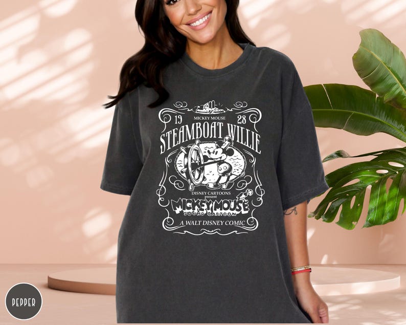 May include: Dark gray t-shirt featuring a white graphic of Mickey Mouse from the 1928 cartoon "Steamboat Willie." The design includes the text "Disney Cartoons" and "A Walt Disney Comic."
