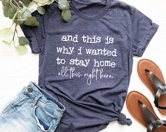May include: Heather navy blue t-shirt with the text "and this is why I wanted to stay home all this right here." The shirt is paired with blue jeans and brown sandals. Green leaves are in the background.