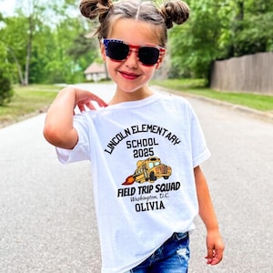 May include: A young girl wearing a white t-shirt with a school bus graphic and the text "Lincoln Elementary School 2025 Field Trip Squad Washington, D.C. Olivia." She is wearing sunglasses with red, white, and blue frames.