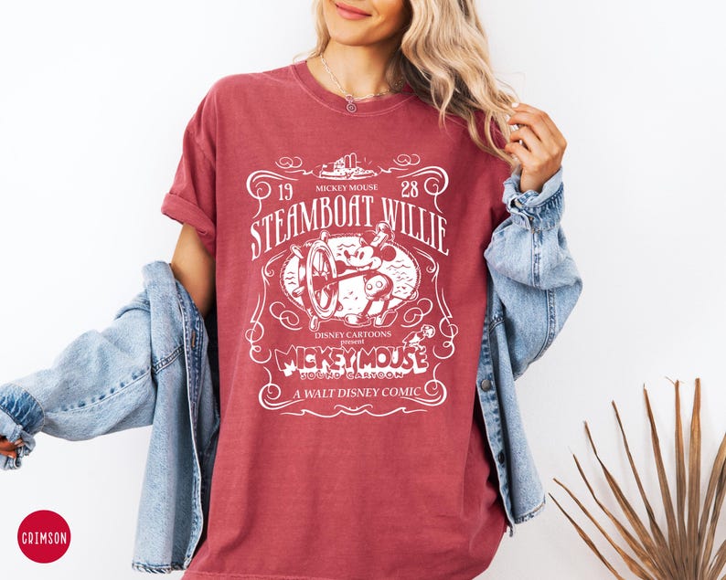 May include: A maroon t-shirt featuring a white graphic of Mickey Mouse from the 1928 Disney cartoon "Steamboat Willie." The design includes the text "Mickey Mouse" and "A Walt Disney Comic."