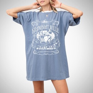 May include: A blue t-shirt dress featuring a white graphic of Mickey Mouse from Steamboat Willie. The graphic includes the text "STEAMBOAT WILLIE", "MICKEY MOUSE", and "A WALT DISNEY COMIC". The dress has short sleeves and a relaxed fit.