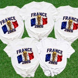 May include: Five white t-shirts featuring a graphic print with the word FRANCE in blue, a gold trophy, the French flag in blue, white, and red, and the year 2026 in blue. Each shirt has a round neckline and short sleeves with a knotted hem detail.