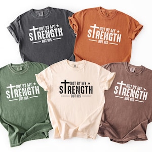 May include: Five t-shirts in various colors: charcoal, rust, olive green, cream, and brown. Each shirt features the text "NOT BY MY STRENGTH BUT HIS" in white, with a cross design. The shirts are laid flat.