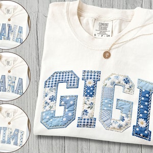 May include: Ivory-colored t-shirt with the name "GIGI" appliqued in various blue patterned fabrics. The letters are outlined with a contrasting stitch. A gold necklace with a pendant is draped on the shirt. Additional shirts with "MAMA", "NANA", and "MIMI" are shown.