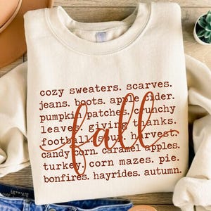 May include: A sand-colored crewneck sweatshirt featuring dark brown text listing fall-themed items like cozy sweaters, scarves, jeans, boots, apple cider, pumpkin patches, crunchy leaves, giving thanks, football, soup, harvest, candy corn, caramel apples, turkey, corn mazes, pie, bonfires, hayrides, and autumn.