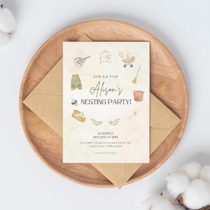 May include: A baby shower invitation with the text "Alison's Nesting Party!" is displayed on a wooden plate with a brown envelope. The invitation features watercolor illustrations of baby-related items, including a stroller and a basket.