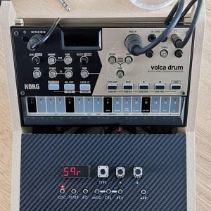Può includere: Un sintetizzatore di percussioni digitali Korg Volca Drum in una cornice di legno. Il sintetizzatore ha vari pulsanti, manopole e un display digitale che mostra "59r". L'immagine include anche un piccolo mixer audio, un bicchiere d'acqua e un rametto di verde.