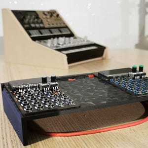 May include: A black electronic music synthesizer with multiple knobs, buttons, and a patterned surface. The synthesizer has a blue side and a red cable. Another synthesizer is in the background, set in a light-colored wooden frame.