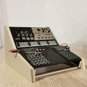Puede incluir: Un sintetizador Korg Volca Drum y otros equipos de música electrónica están montados en un soporte de madera blanco. El sintetizador tiene paneles negros y plateados con perillas y teclas. Cables rojos conectan los componentes.
