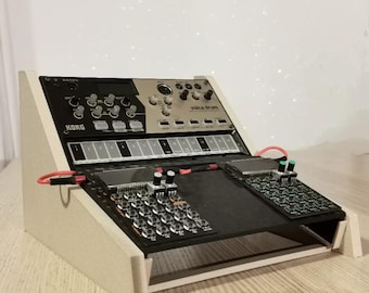 Könnte beinhalten: Ein Korg Volca Drum Synthesizer und andere elektronische Musikgeräte sind auf einem weißen Holzständer montiert. Der Synthesizer hat schwarze und silberne Paneele mit Knöpfen und Tasten. Rote Kabel verbinden die Komponenten.