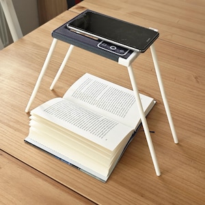 Könnte beinhalten: Ein schwarzes Smartphone liegt auf einem marineblauen und weißen Ständer mit weißen Beinen, über einem offenen Buch auf einem Holztisch. Der Ständer ist für ein Telefon oder Tablet konzipiert.