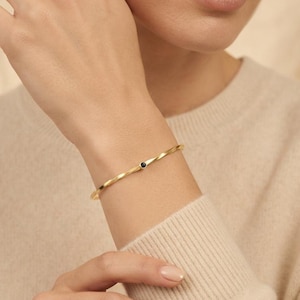 Op de afbeelding: Een dunne gouden armband met een gedraaid metalen bandontwerp en een enkele kleine, ronde, zwarte edelsteen in het midden. De armband heeft een gepolijste afwerking en een delicate, minimalistische structuur die om de pols sluit.