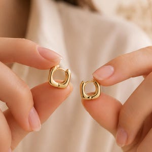 May include: Two gold-colored square hoop earrings are held up in front of a soft, neutral background. The earrings have a polished finish and a simple, modern design. The earrings are small and appear to be lightweight.