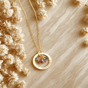 May include: A gold necklace featuring a circular pendant with the names "Alex" and "Emily" engraved. The pendant holds several gemstones in various colours. The necklace is displayed with white flowers.