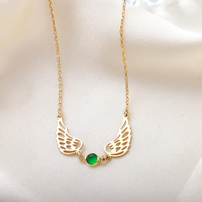 Pode incluir: Um colar dourado com um par de asas estilizadas em ambos os lados de uma pequena pedra preciosa verde redonda. O colar tem uma corrente delicada e &eacute; exibido em um fundo branco.