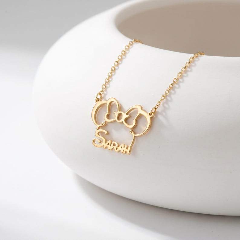 May include: A gold-toned necklace featuring a pendant shaped like a stylized mouse head with a bow, incorporating the name Sarah in a cursive script font. The pendant is attached to a delicate gold-toned chain with small, uniform circular links.