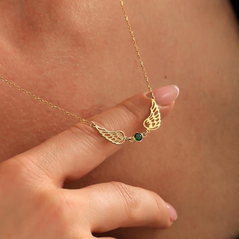 Pode incluir: Um colar dourado com um design de asas e uma pedra preciosa verde. As asas est&atilde;o em ambos os lados da pedra preciosa. O colar &eacute; usado em volta do pesco&ccedil;o. As asas t&ecirc;m padr&otilde;es recortados detalhados.