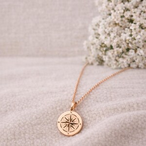 May include: A rose gold-toned circular pendant necklace featuring an engraved compass rose design. The pendant is suspended from a matching delicate chain, with the cardinal directions N, E, S, and W clearly marked on the face of the metal disc.