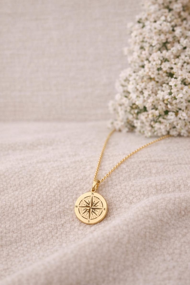 May include: A gold-toned circular pendant necklace featuring an engraved compass rose design with the cardinal directions N, E, S, and W clearly marked. The pendant hangs from a delicate gold-toned chain, showcasing a polished metallic finish.