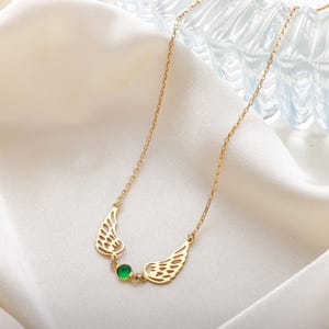 Pode incluir: Um colar dourado com um par de asas estilizadas em cada lado de uma pequena pedra preciosa verde redonda. As asas t&ecirc;m um design vazado detalhado. O colar &eacute; exibido em um tecido branco e sedoso.