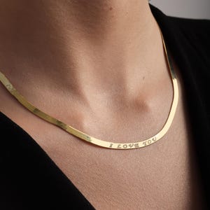 Könnte beinhalten: Eine goldfarbene Fischgräten-Halskette mit flachem, flexiblem Design. Die Oberfläche der Kette ist mit dem Text "I LOVE YOU" in Großbuchstaben graviert, der entlang der unteren Kurve der Kette positioniert ist, während sie am Hals anliegt.