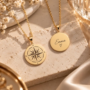 May include: Two gold-toned necklaces with circular pendants. One pendant features a detailed black compass rose engraving with cardinal directions. The other pendant is engraved with the name Emma in cursive script above a small heart symbol.