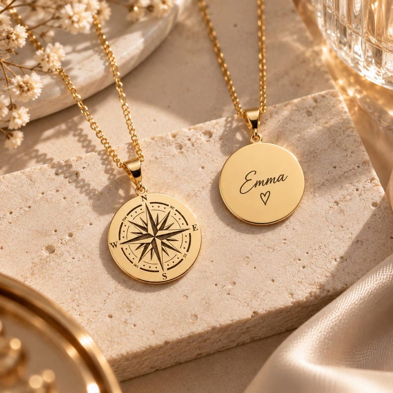 May include: Two gold-toned necklaces with circular pendants. One pendant features a detailed black compass rose engraving with cardinal directions. The other pendant is engraved with the name Emma in cursive script above a small heart symbol.