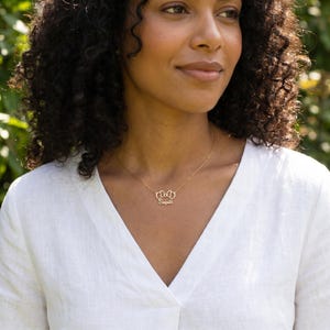 May include: A gold-toned necklace featuring a delicate chain and a central pendant shaped like a bow with the name Sophia written in cursive script. The pendant is positioned at the center of the collarbone area on a white linen shirt.