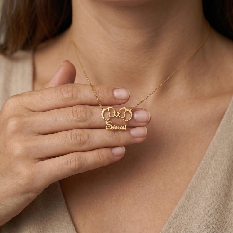 May include: A gold-toned necklace featuring a pendant shaped like a mouse head silhouette with a bow at the top and the name Sarah written in a cursive script font integrated into the lower portion of the design on a thin metal chain.