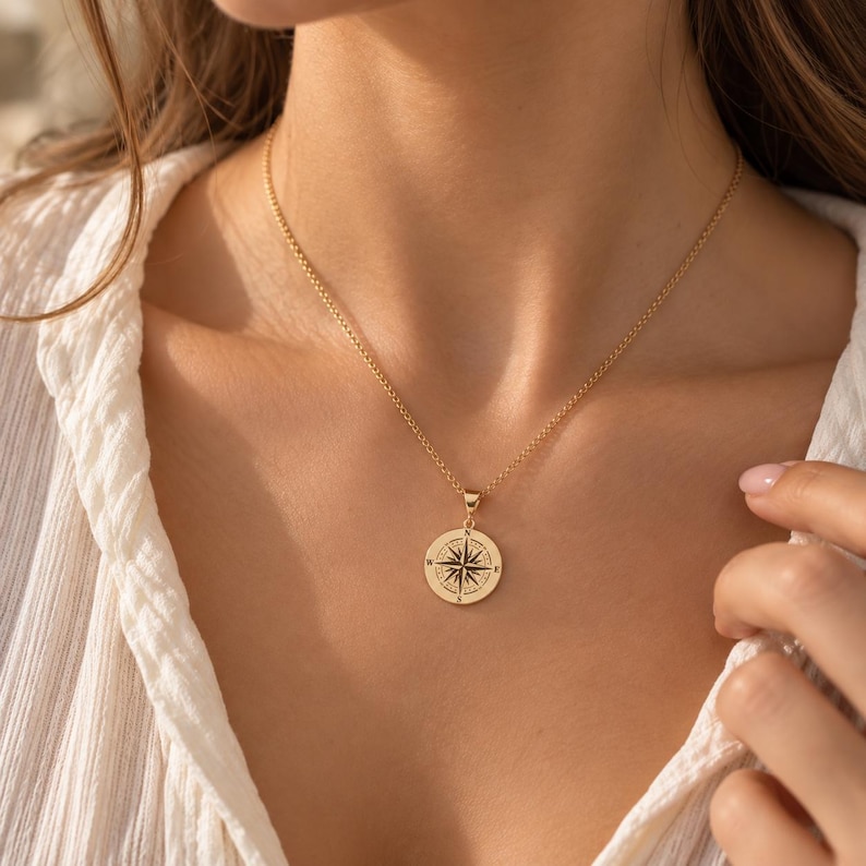 May include: A gold-toned necklace featuring a circular pendant with a black engraved compass rose design. The pendant hangs from a delicate gold-toned chain. The compass rose includes cardinal direction letters N, S, E, and W clearly visible.