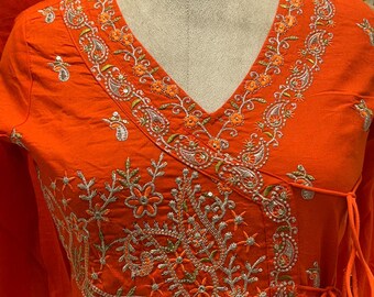 May include: Bright orange wrap-style tunic with intricate embroidery around the V-neck and bodice. The embroidery features floral and paisley designs in shades of orange, silver, and green. The tunic has long sleeves and a gathered skirt.