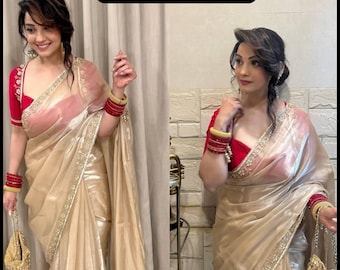 May include: A woman wearing a gold saree with a red blouse. The saree has a shimmering effect and is adorned with a delicate embroidered border. The woman is also wearing red and gold bangles and carrying a gold purse. The text "BoutiquebyRihanaa" is at the top.