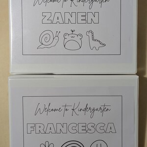 May include: Two white binders with custom covers. The top binder says "Welcome to Kindergarten" and "ZANEN" in a bold font, with illustrations of a snail, frog, and dinosaur. The bottom binder says "FRANCESCA" with a flower, rainbow, and smiley face.