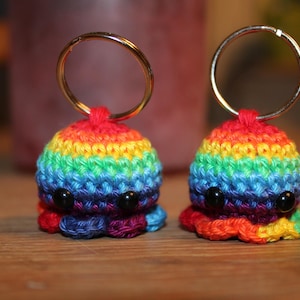 Könnte beinhalten: Zwei handgefertigte Häkel-Oktopus-Schlüsselanhänger. Jeder Oktopus ist aus lebendigem, regenbogenfarbenem Garn gefertigt und hat schwarze Perlenaugen. Die Schlüsselanhänger haben goldfarbene Metallringe oben befestigt. Die Oktopusse sitzen auf einer Holzoberfläche.