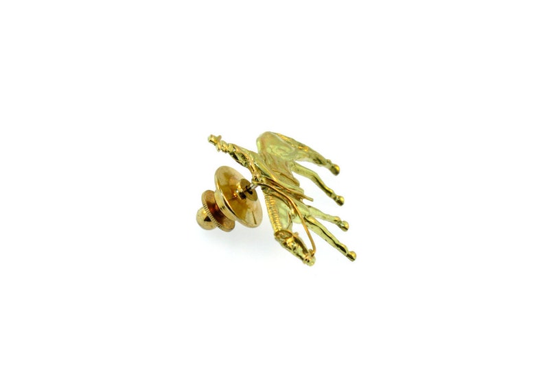 May include: A gold-toned pin featuring a horse and rider. The pin has a safety clasp on the back.