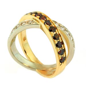May include: A two-tone gold and silver ring with an intertwined design. One band is gold and set with a row of dark red gemstones. The other band is silver and set with small, clear gemstones. The ring is on a white background.
