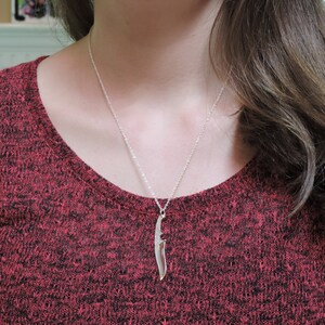 May include: A silver necklace with a knife pendant. The necklace is worn by a person with long brown hair wearing a red sweater.