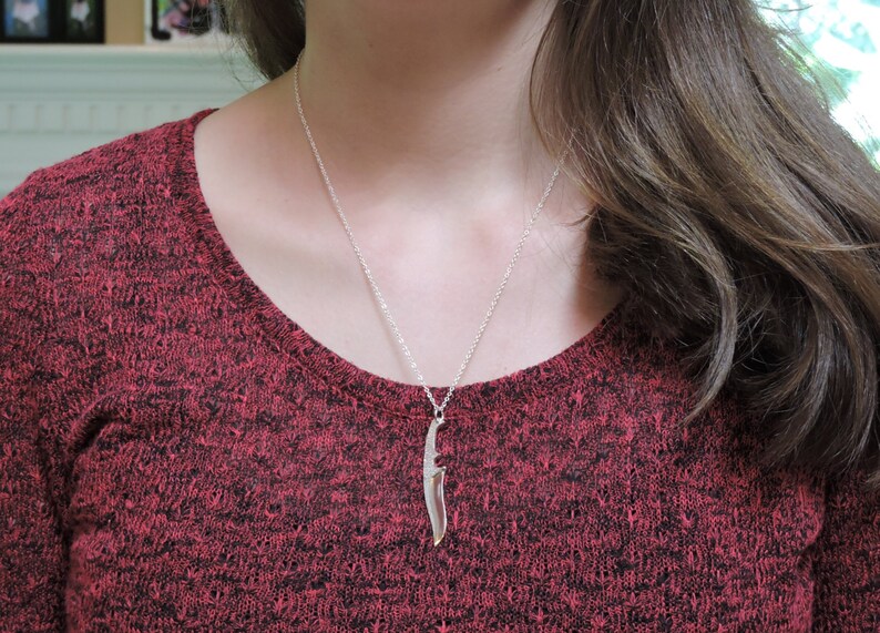 May include: A silver necklace with a knife pendant. The necklace is worn by a person with long brown hair wearing a red sweater.