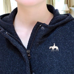 May include: A dark gray hooded sweatshirt with a zipper closure and a small gold pin of a rider on a horse.