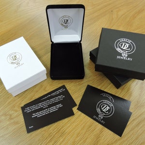 May include: Three black boxes with the logo "Leslie Jewelry" and a white circle with the letter "L" inside. The boxes are stacked on top of each other. There is also a white box with the same logo and a card with the same logo and the text "Leslie Jewelry is composed of the original handmade jewelry of Diana and Raymond, both born in Leslie, Scotland. They continued this tradition as long as we will repair and refinish their work. We take great pride in making original handmade jewelry and never compromise on that promise. Thank you for your advertisement."