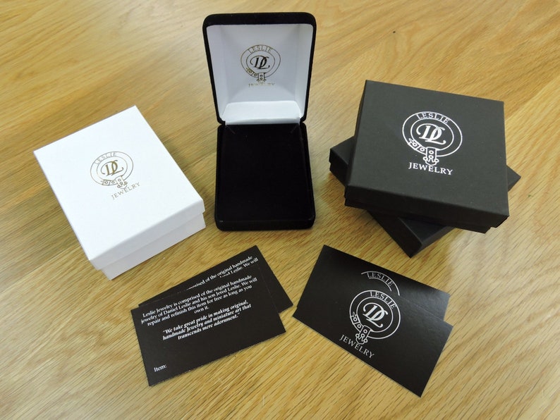 May include: Three black boxes with the logo "Leslie Jewelry" and a white circle with the letter "L" inside. The boxes are stacked on top of each other. There is also a white box with the same logo and a card with the same logo and the text "Leslie Jewelry is composed of the original handmade jewelry of Diana and Raymond, both born in Leslie, Scotland. They continued this tradition as long as we will repair and refinish their work. We take great pride in making original handmade jewelry and never compromise on that promise. Thank you for your advertisement."