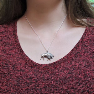 May include: A silver necklace with a detailed buffalo pendant. The necklace is worn by a person wearing a red sweater.