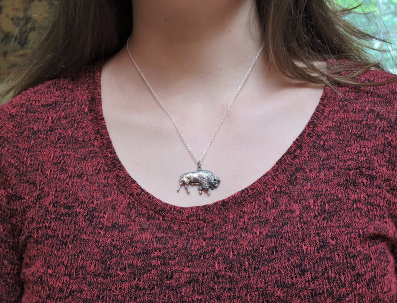 May include: A silver necklace with a detailed buffalo pendant. The necklace is worn by a person wearing a red sweater.