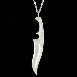 May include: A silver pendant necklace featuring a stylized knife design. The pendant has a textured surface and a curved shape.
