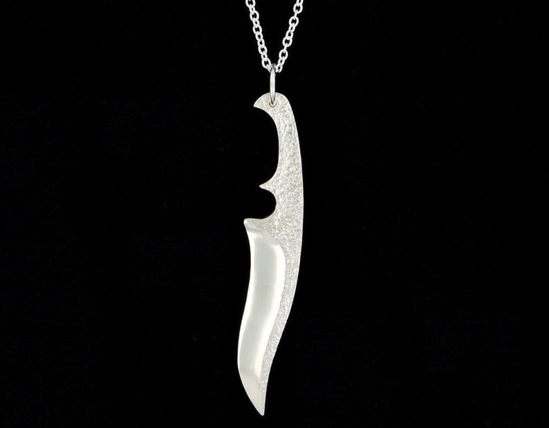 May include: A silver pendant necklace featuring a stylized knife design. The pendant has a textured surface and a curved shape.