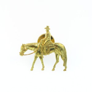 May include: A gold-colored metal pin depicting a rider on a horse. The rider is wearing a hat and is sitting in a saddle. The horse is in a walking position.