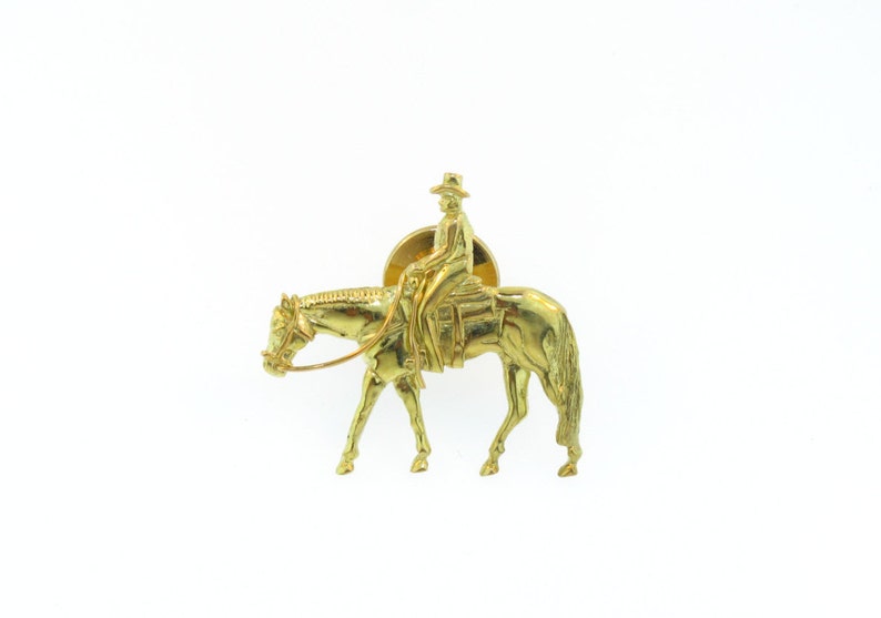 May include: A gold-colored metal pin depicting a rider on a horse. The rider is wearing a hat and is sitting in a saddle. The horse is in a walking position.